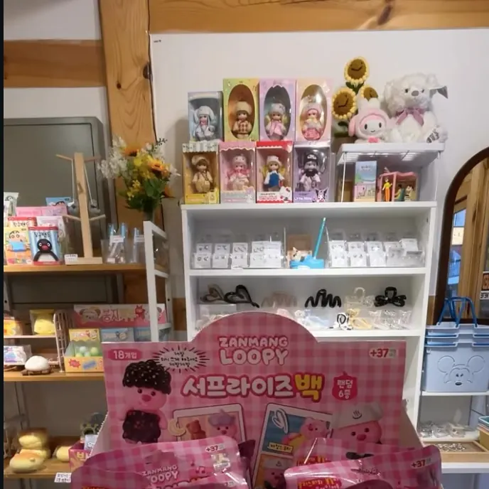 나무 기둥 옆 흰 선반에 상자 인형과 헤어 액세서리가 놓여 있고, 앞에 랜덤 상품 진열대가 있는 매장 안